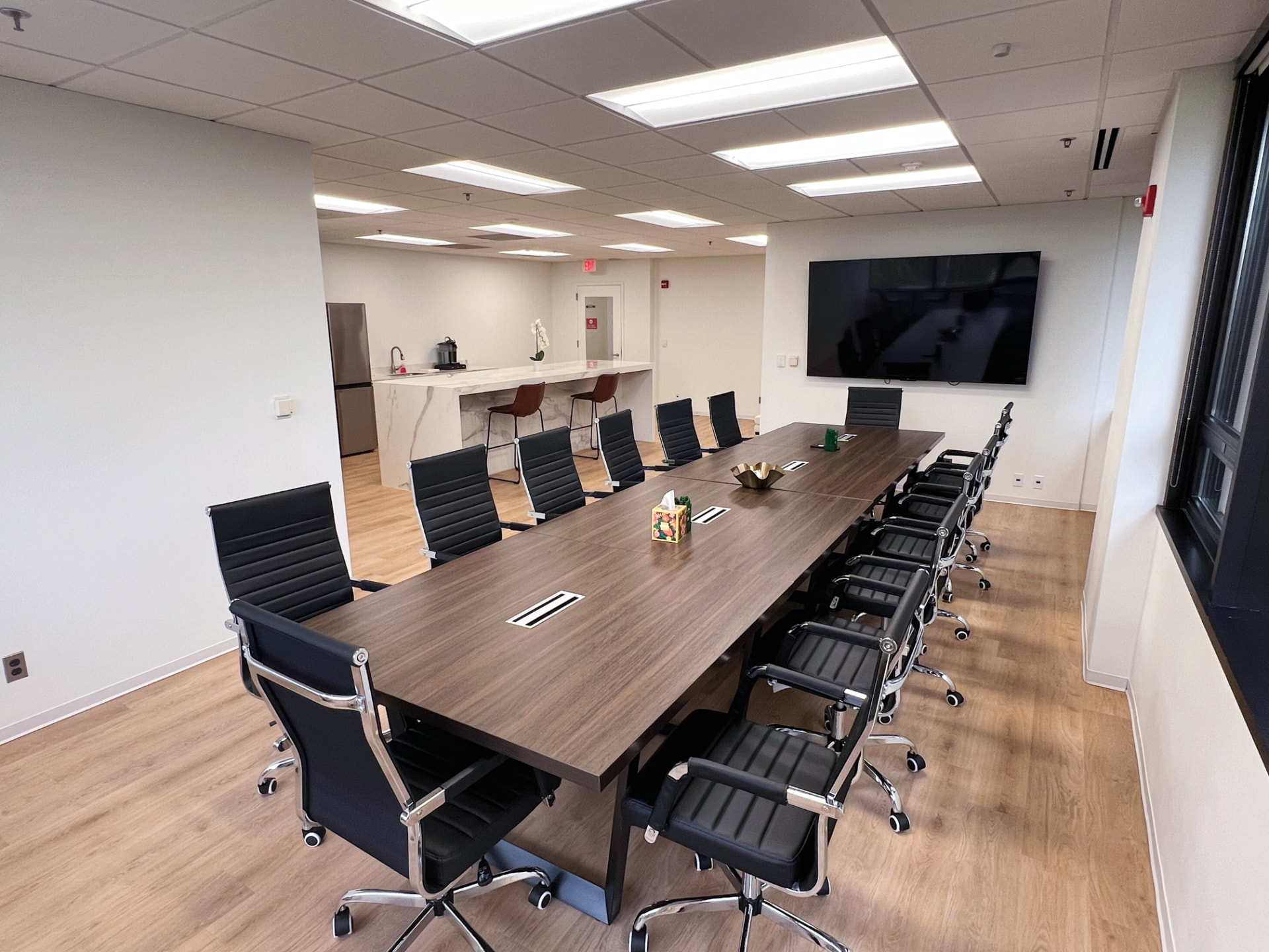 Modern conference room with a long executive table, black leather chairs, wall-mounted screen, and kitchenette area